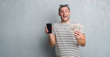 Dolar ve smarphone çok mutlu ve heyecanlı, kazanan ifade ile çığlık zaferini kutluyor büyük holding gri grunge duvarın üzerinden beyaz delikanlı gülümse ve el kaldırdı