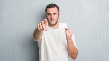 Beyaz delikanlı boş ziyaret kartı kamera ve size parmak ile işaret gösteren gri grunge duvarın üzerinden, el işareti, pozitif ve kendinden emin bir jest ön