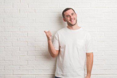 Genç beyaz adam arıyor ve başparmak yanında yukarıyı mutlu yüz ile gülümseyen beyaz tuğla duvarın üzerinden ayakta.
