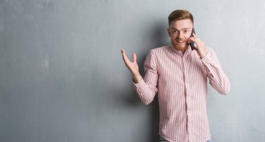 Genç Kızıl saçlı adam gri grunge duvar büyük gülümseme ile çığlık zaferini kutluyor ve ellerini kaldırdı telefon çok mutlu ve heyecanlı, kazanan ifade üzerinde konuşuyor