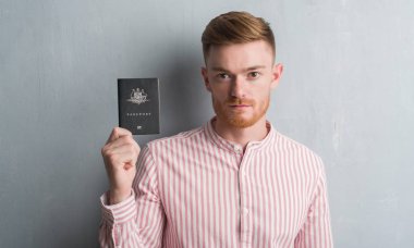 Young redhead man over grey grunge wall holding passport of Australia with a confident expression on smart face thinking serious clipart