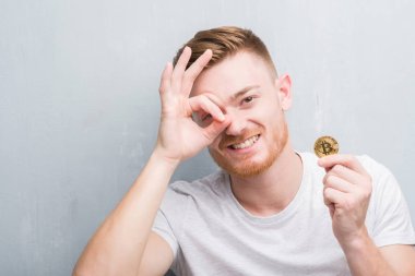 Gri grunge Duvar ile mutlu yüz el parmaklarının arasından seyir göz üzerinde ok işaretiyle yapıyor gülümseyen bitcoin gösteren genç Kızıl saçlı adam