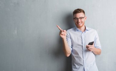 Genç Kızıl saçlı adam gri grunge duvar el ve parmak ile tarafına işaret çok mutlu smartphone kullanarak ileti gönderme