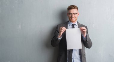 Genç redhead iş adamı boş kağıt sözleşme ile ayakta ve diş gösterilen kendine güvenen bir gülümsemeyle gülümseyerek mutlu bir yüz tutan gri grunge duvarın üzerinden