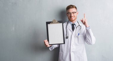 Genç redhead Doktor adam gri grunge holding Pano bir fikirle sürpriz duvar veya gülen yüz, bir numara ile işaret parmağı soru