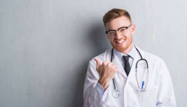 Genç redhead Doktor adam işaret ve başparmak kadar mutlu yüz gülümseyen tarafı ile gösterilen gri grunge duvarın üzerinden