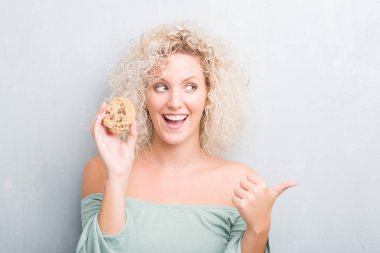 Sarışın genç kadın işaret ve başparmak kadar mutlu yüz gülümseyen tarafı ile gösterilen çikolatalı bisküvi yemek grunge gri duvarın üzerinden