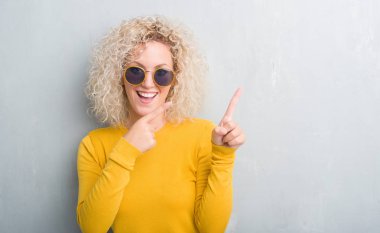 Grunge üzerinde Kıvırcık saçlı sarışın genç kadın gülümseyerek ve iki elleri ve parmakları ile tarafına işaret kamera bakarak arka plan gri.