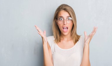 Grunge gri üzerinde duran güzel genç kadın duvar büyük gülümseme ile çığlık ve ellerini kaldırdı giyen gözlük çok mutlu ve heyecanlı, kazanan ifade kutluyor zafer