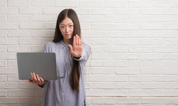年轻的中国妇女在砖墙使用手提电脑与张开手做停止标志以严肃和自信的表达 防御手势 — 图库照片