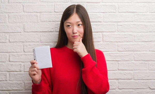 年轻的中国妇女在砖墙上显示空白笔记本严肃的面孔思考问题 非常迷惑想法 — 图库照片