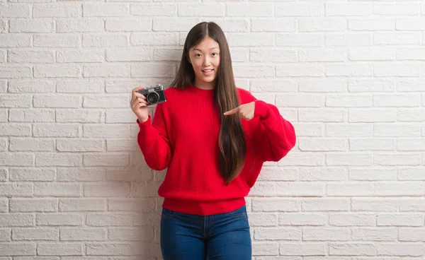 Young Chinese woman over brick wall holding vintage camera with surprise face pointing finger to himself