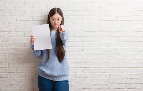 Genç Çinli kadın kamera ve size parmak ile işaret boş kağıt tutan tuğla duvarın üzerinden el işareti, pozitif ve kendinden emin bir jest ön