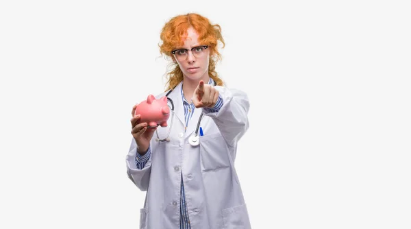 Young Redhead Doctor Woman Holding Piggy Bank Pointing Finger Camera — Stock Photo, Image