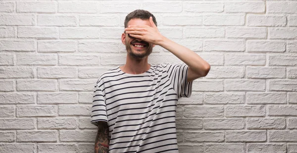 Genç yetişkin adam gülümsüyor ve gözleri için sürpriz kapsayan yüz el ile gülüyor beyaz tuğla duvarın üzerinden duran güneş gözlüğü takıyor. Kör kavramı.