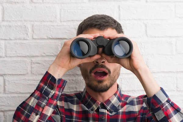 Genç yetişkin adam dürbün bir sürpriz yüz, korkuyor ve heyecanlı korku ifadesi ile şok korkmuş üzerinden seyir tuğla duvarın üzerinden