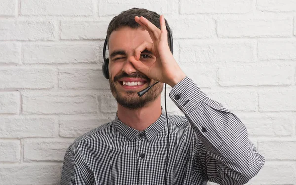 Young adult man over brick wall working as operator with happy face smiling doing ok sign with hand on eye looking through fingers