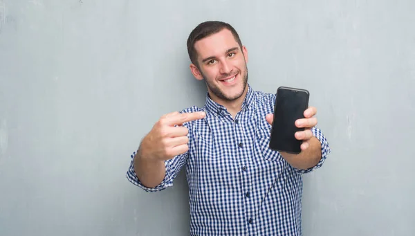 年轻的白种人在灰色的垃圾墙上显示手机屏幕的空白 用手和手指很开心的指向 — 图库照片