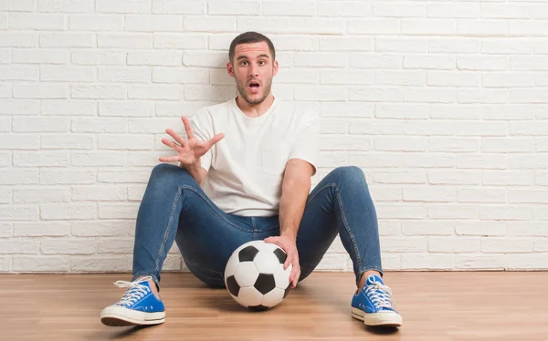 驚き顔 恐怖表現と興奮とショックで怖いサッカー サッカー ボールを保持している白いレンガの壁に座っている若い白人男 — ストック写真