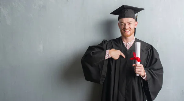 自分自身に驚き顔ポインティング指で学位を保持研究科の制服を着て灰色グランジ壁を越えて若い赤毛の男 — ストック写真