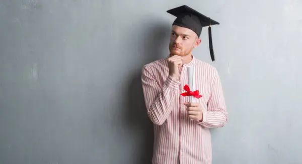 灰色のグランジの赤毛の若い男壁程度深刻な顔を考えて質問 非常に混乱して考えを保持している大学院の帽子を着用 — ストック写真