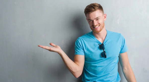Young redhead man over grey grunge wall wearing casual outfit smiling cheerful presenting and pointing with palm of hand looking at the camera.