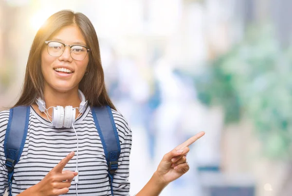 笑顔で側に つの手と指で指しているカメラを見て孤立の背景にヘッドフォンとバックパックを着ている若いアジア学生女性 — ストック写真