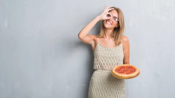 指の間から見て目に手で サインをして笑っている幸せそうな顔でペパロニのピザを食べてグランジ灰色の壁を越えて美しい若い女性 — ストック写真