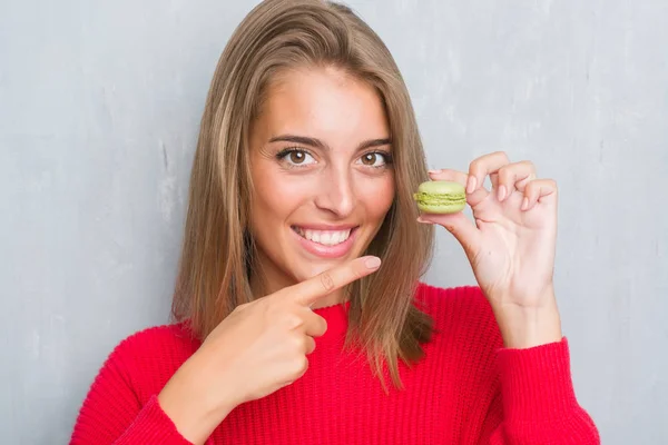 緑マカロン手と指で指している非常に幸せを食べるグランジ灰色の壁に美しい若い女性 — ストック写真