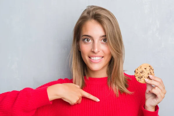 グランジに美しい若い女性灰色の壁が指を指す自身驚き顔でチョコレート チップ クッキーを食べて — ストック写真