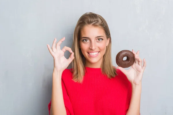 優秀なシンボルの指で サインをしているチョコレートのドーナツを食べてグランジ灰色の壁に美しい若い女性 — ストック写真