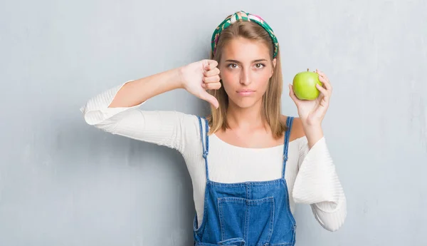 美丽的年轻妇女在垃圾灰色墙壁吃绿色苹果与愤怒的面孔 消极标志显示不喜欢与拇指下来 拒绝概念 — 图库照片