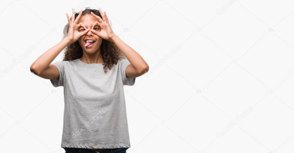 Beautiful young hispanic woman wearing glasses doing ok gesture like binoculars sticking tongue out, eyes looking through fingers. Crazy expression.