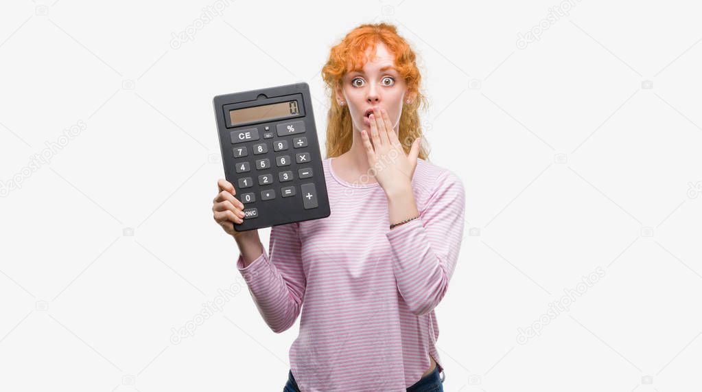 Young redhead woman holding big calculator cover mouth with hand shocked with shame for mistake, expression of fear, scared in silence, secret concept