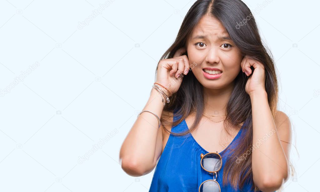 Young asian woman over isolated background covering ears with fingers with annoyed expression for the noise of loud music. Deaf concept.
