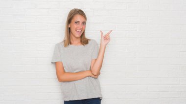 Güzel ve genç birbayan yüzünde kocaman bir gülümseme ile beyaz tuğla duvarın üzerinden kameraya bakarak yan el ve parmak ile işaret.
