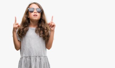 Esmer Latin kız güneş gözlüğü takıyor hayrete ve yukarıya bakıyor ve parmakları ile işaret ve silah kaldırdı şaşırttı.
