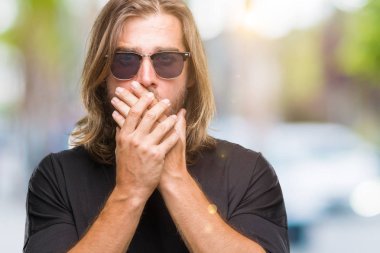 Üzerinde güneş gözlüğü takmış uzun saçlı Genç yakışıklı adam arka plan ağız hata için elleriyle kapsayan şok izole. Gizli kavramı.