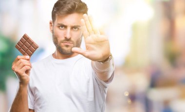 Yakışıklı delikanlı ile izole arka plan üzerinde çikolata yeme dur işareti ile ciddi ve kendine güvenen ifade, savunma hareketi yaparken el açmak