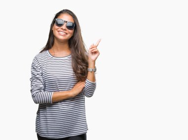 Kameraya bakarak yan el ve parmak ile işaret yüzünde kocaman bir gülümseme ile izole arka plan üzerinde güneş gözlüğü takmış genç güzel Arap kadın.