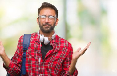 Üzerinde kulaklık ve sırt çantası giyiyor yetişkin İspanyol öğrenci adam arka plan clueless ve şaşkın bir ifadeyle kollar ve eller kaldırdı izole. Şüphe kavramı.