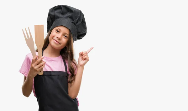黑发西班牙裔女孩穿着厨师制服非常高兴地指着手和手指的侧面 — 图库照片