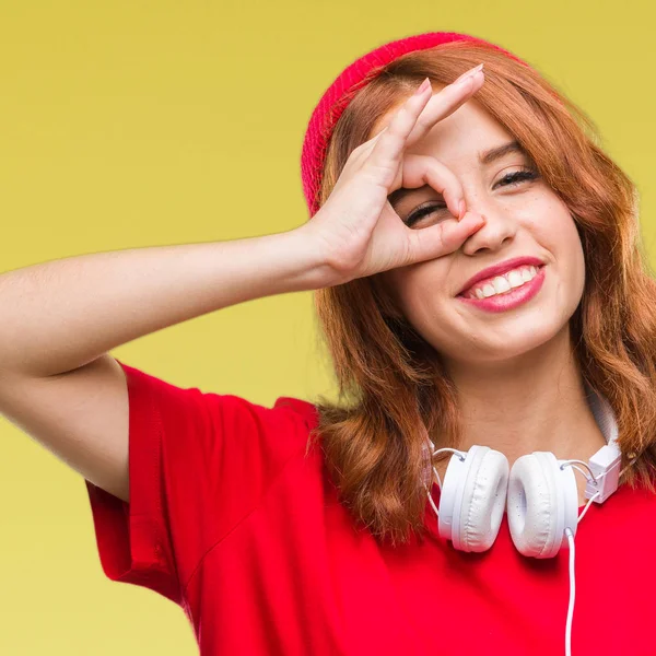 İzole arka plan kulaklık ve gülümseyen, parmakları ile mutlu yüz üzerinden seyir göz el ile ok jest yapıyor kap giyen genç güzel hipster kadın.