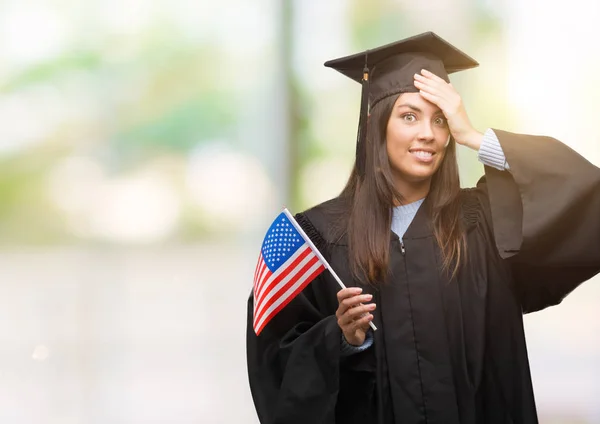Young hispanic woman wearing graduated uniform holding flag of america stressed with hand on head, shocked with shame and surprise face, angry and frustrated. Fear and upset for mistake.