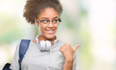 Kulaklıklar ve sırt çantası arıyorum ve başparmak yanında işaret mutlu yüz ile gülümseyen izole arka plan üzerinde giyen genç afro Amerikan öğrenci kadın.