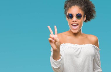 Mutlu yüz zafer işareti yaparken kameraya göz kırpıyor ile gülümseyen izole arka plan üzerinde güneş gözlüğü takmış genç afro Amerikalı kadın. İki numaralı.