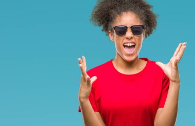 Genç afro Amerikalı kadın deli ve silah ile başarı için deli kutluyor izole arka plan üzerinde güneş gözlüğü takıyor kaldırdı ve heyecanlı çığlık gözler kapalı. Kazanan kavramı