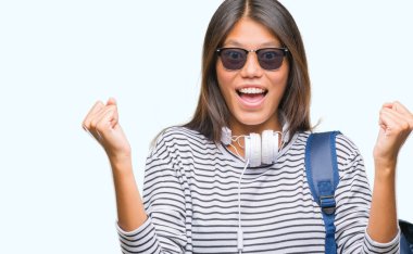 Üzerinde kulaklık ve sırt çantası giyen genç Asyalı öğrenci kadın şaşırmış ve hayret için başarı ile silah kaldırdı kutluyor arka plan izole ve gözleri açık. Kazanan kavramı.