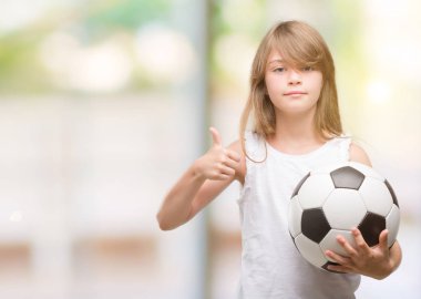 Genç Sarışın yürümeye başlayan çocuk futbol topu ok işareti, başparmak ile parmak, mükemmel işareti yapıyor kocaman bir gülümseme ile mutlu holding