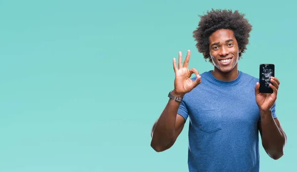 Afro american man holding broken smartphone over isolated background doing ok sign with fingers, excellent symbol